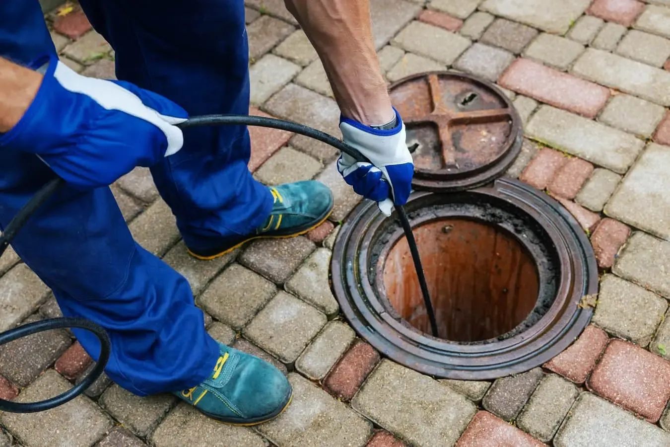 A Drain Medics engineer performing drain unblocking or a CCTV drain survey by inserting a cable into an open manhole on a block-paved driveway.